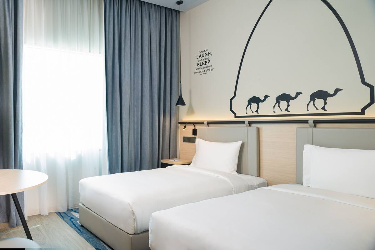 A hotel room with two beds, a small table with apples, a chair, and a wall with a camel silhouette and a quote.