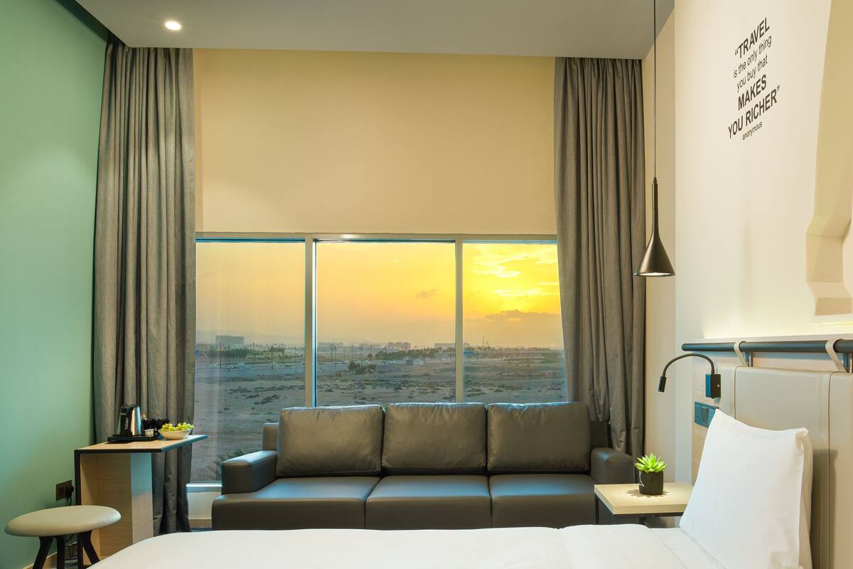 A modern hotel room with a large bed, a sofa, a desk, and a view of the sunset.
