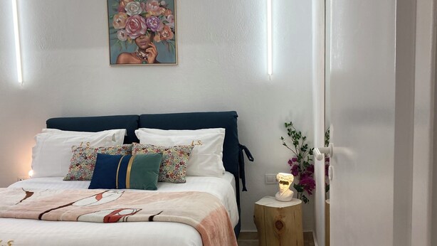 A bedroom with a bed, pillows, a nightstand, a painting, and a vase with flowers. - Lucky Suites (Rethymno)