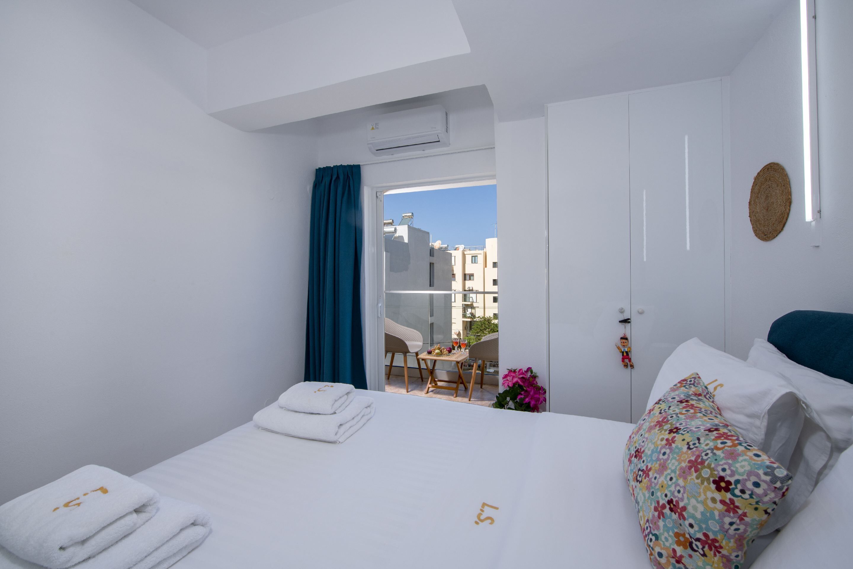 A hotel room with a bed, a small table with a chair, a balcony with a view of buildings, and a door with a keyhole.