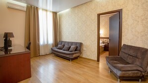 A hotel room with a wooden floor, a sofa, a desk with a lamp, and a bed in the adjacent room. - Park Hotel Tskaltubo Balneo Resort (Tsqaltubo)