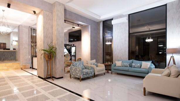 A spacious living area with a large sofa, a smaller armchair, and a coffee table. A potted plant is placed near the entrance, and a chandelier hangs from the ceiling. - Park Hotel Tskaltubo Balneo Resort (Tsqaltubo)