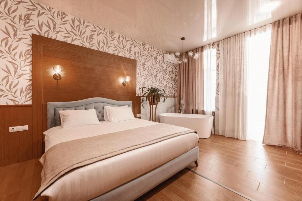 A bedroom with a bed, a wooden headboard, a wall-mounted light fixture, a large window with curtains, and a plant.