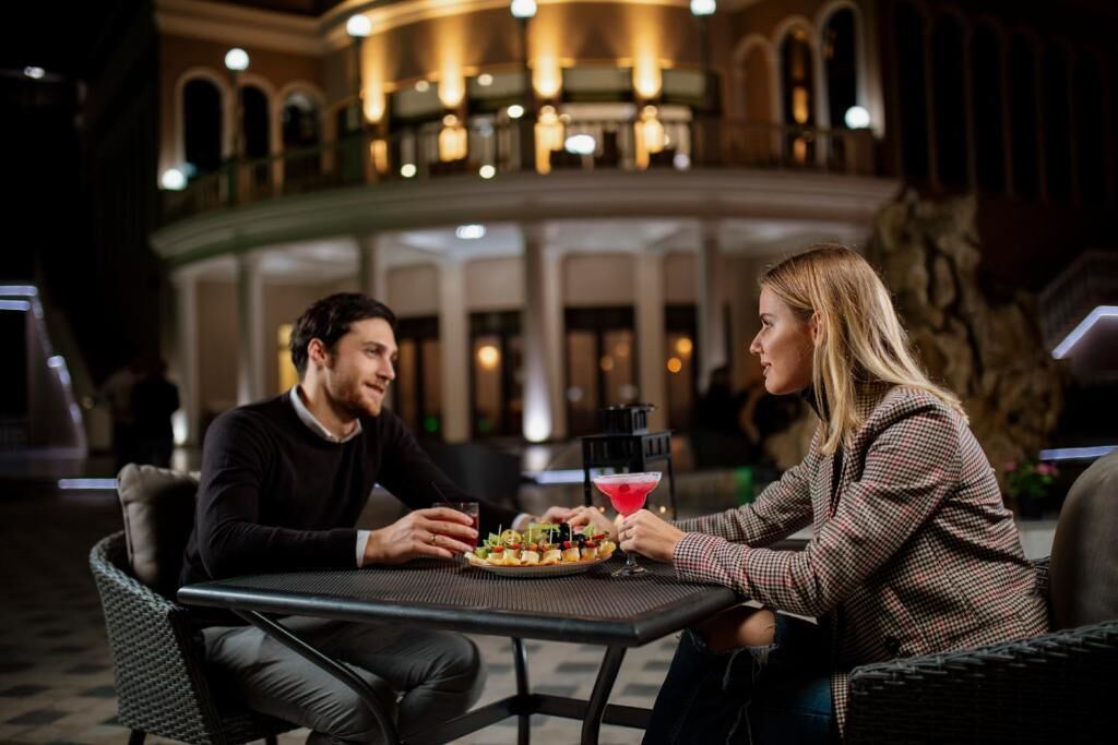 A couple seated at a table with a cocktail and appetizers in a hotel lobby with a grand staircase and arched windows.