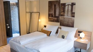 Una cama doble con sábanas blancas, una mesita de noche con lámpara y un cuadro en la pared.