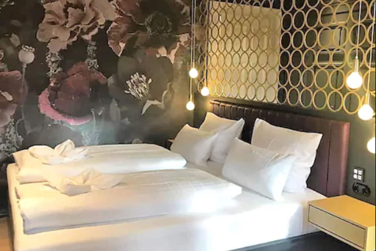 Una cama bien hecha con sábanas y almohadas blancas, un decorativo muro con motivos florales y una lámpara moderna sobre la cama.