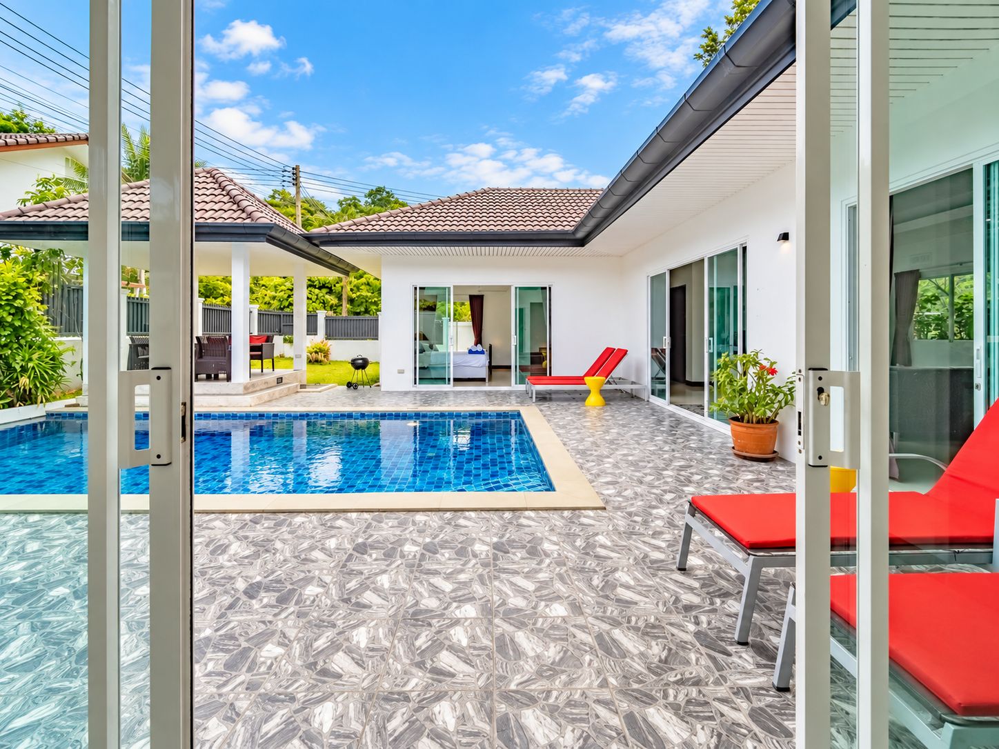 Un área de piscina exterior moderna con solárium empinado, sillas de descanso rojas y una puerta de cristal que da a un porche cubierto.