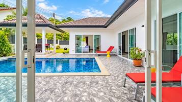 Un área de piscina exterior moderna con solárium empinado, sillas de descanso rojas y una puerta de cristal que da a un porche cubierto.