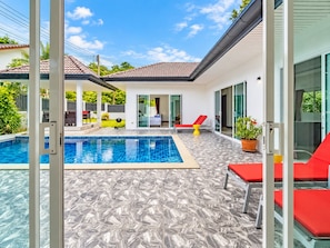 Un área de piscina exterior moderna con solárium empinado, sillas de descanso rojas y una puerta de cristal que da a un porche cubierto.