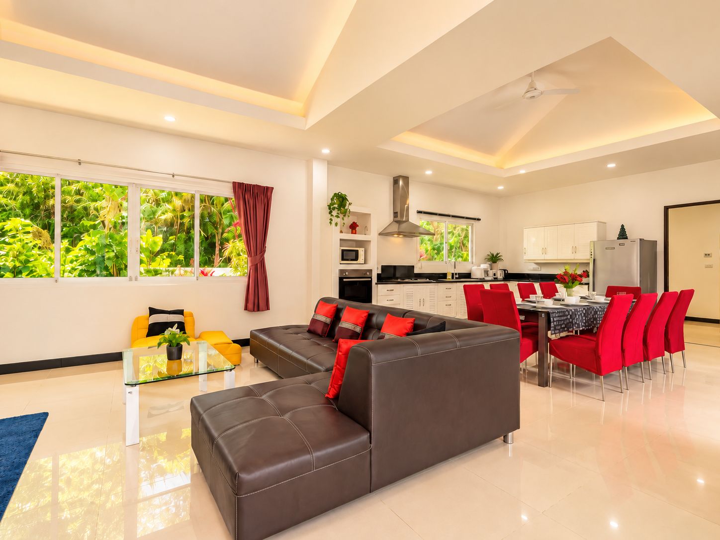 A modern living room with a sofa, a glass coffee table, and a dining area with red chairs.