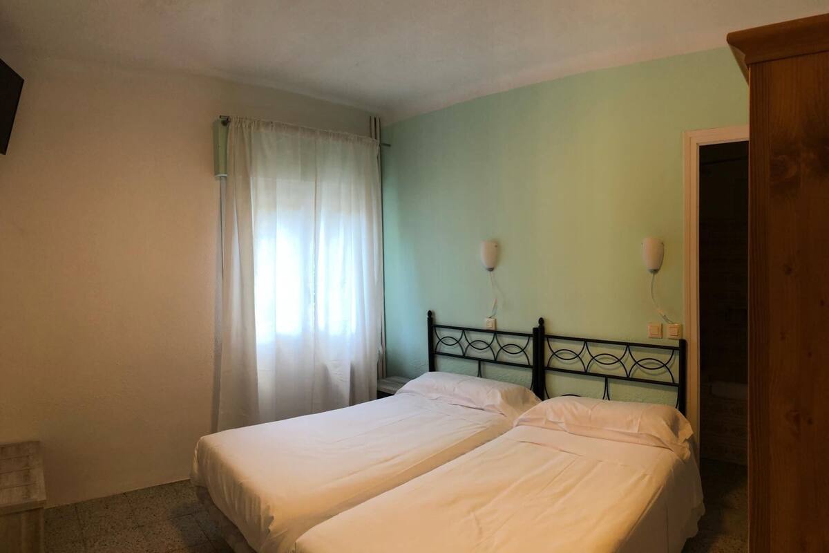 A bedroom with two single beds, a wooden wardrobe, a nightstand with a lamp, and a window with curtains.