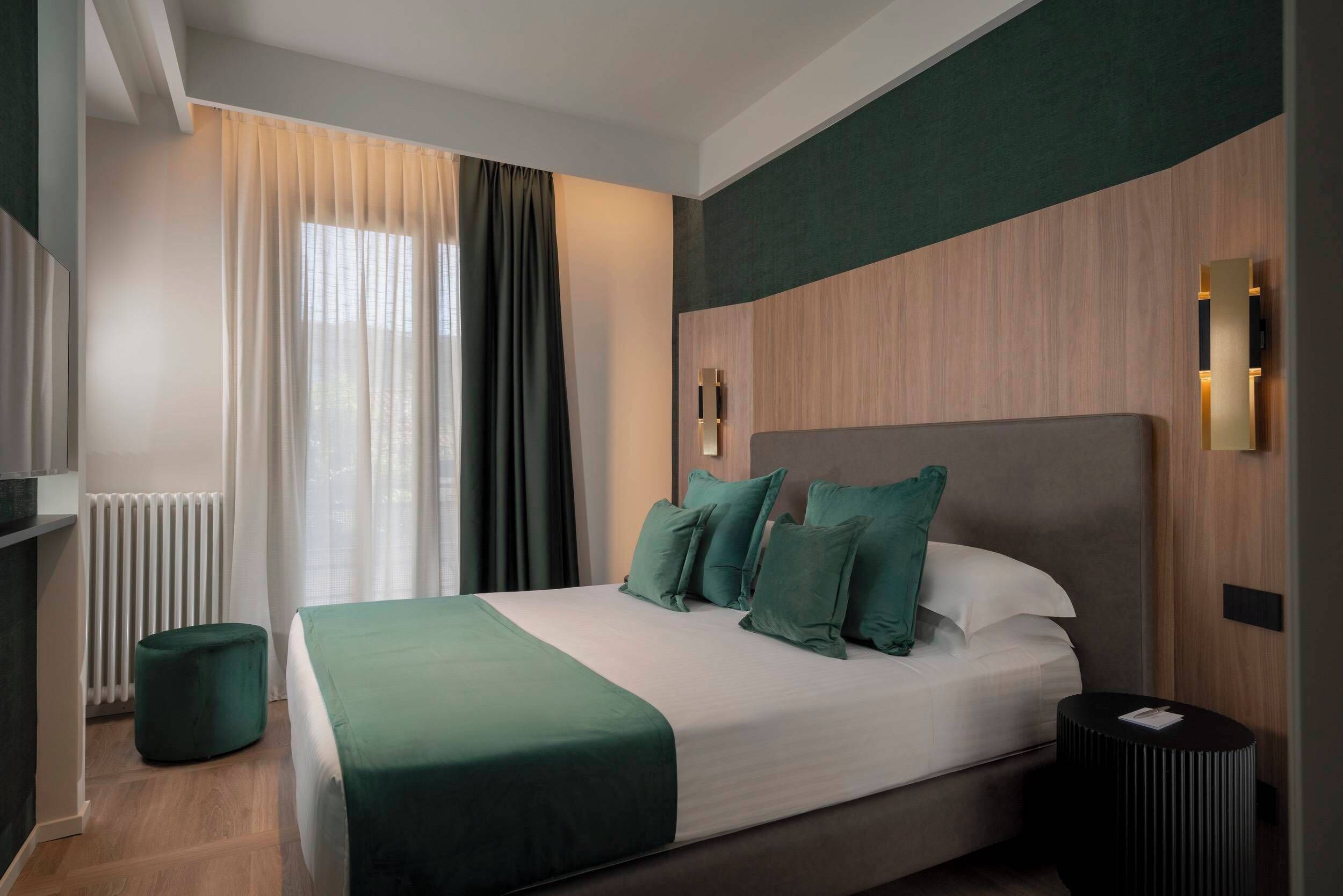 Habitación de hotel moderna con una cama grande, un reposapiés redondo, una mesita de noche y una lámpara fijada a la pared.