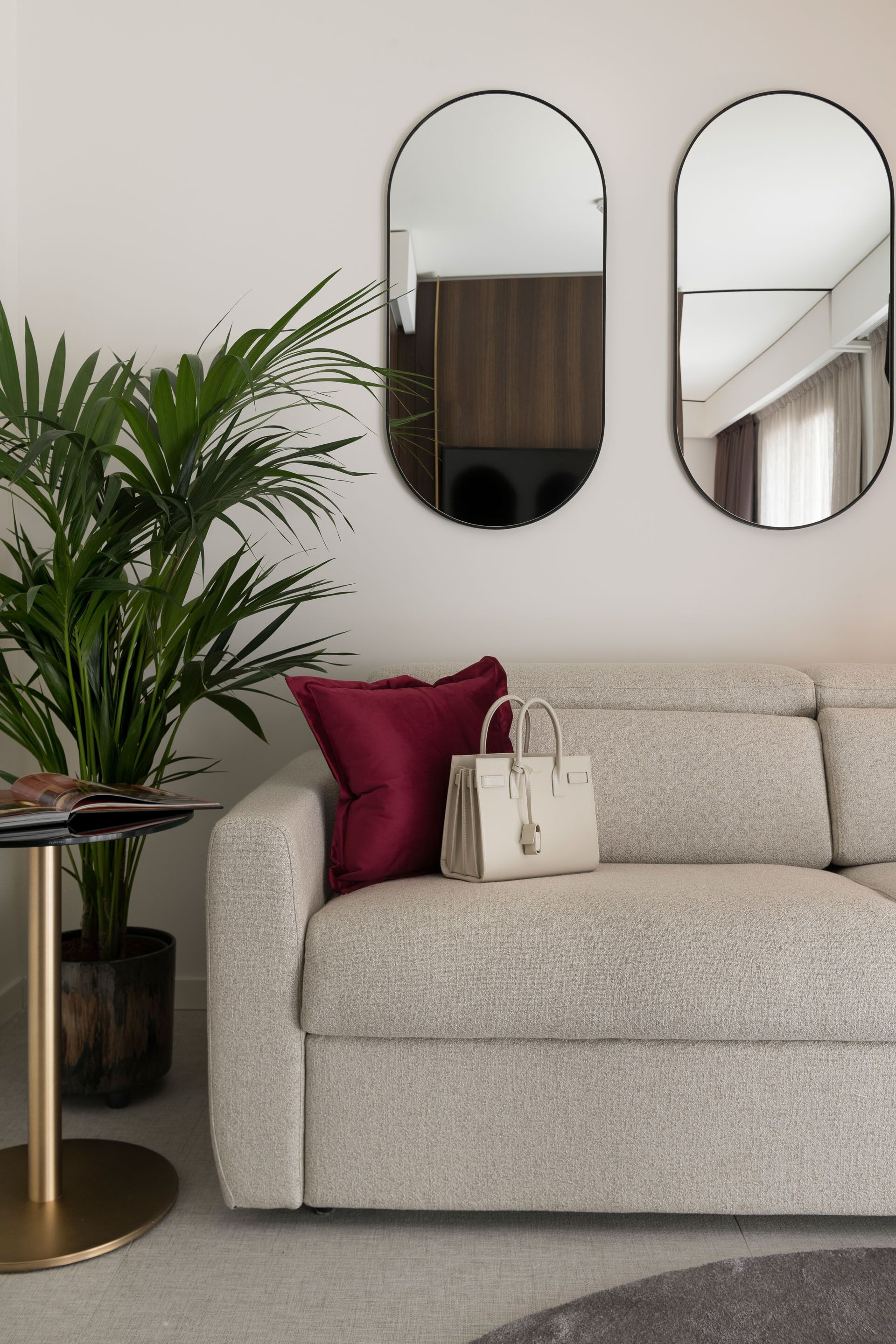 A neutral-toned sofa with a red pillow and a white handbag, a round table with a plant, and two wall-mounted mirrors.