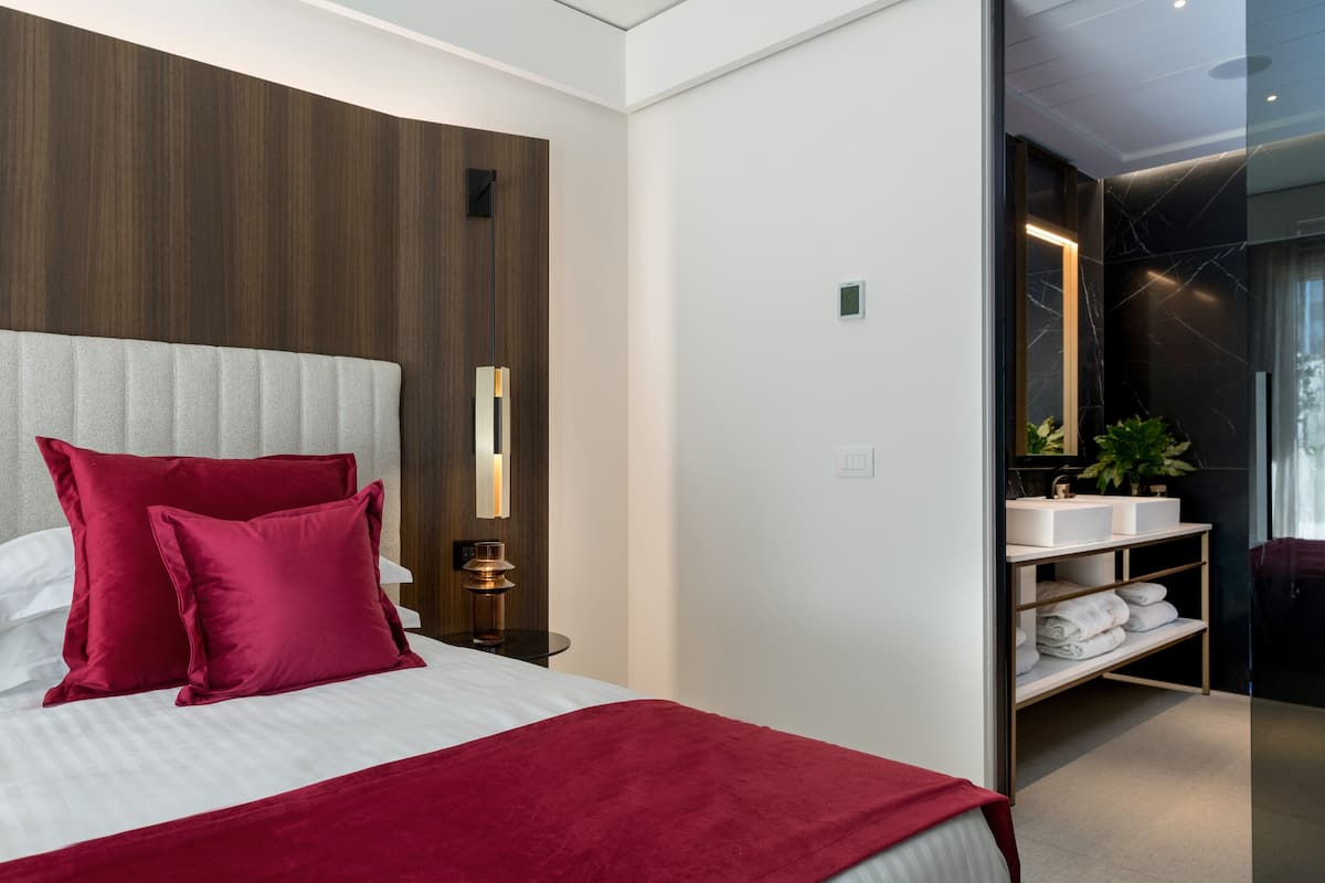 Una habitación de hotel moderna con una cama grande, almohadas rojas, cabecero de madera, una mesita de noche con lámpara, un espejo y un baño con lavamanos y toallero.
