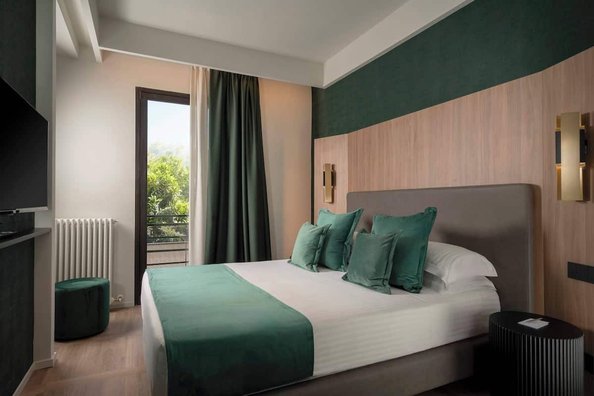 Habitación de hotel moderna con una cama grande, vistas a zonas verdes por una ventana y una pequeña mesa redonda.