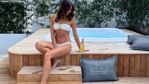 A woman sitting on a wooden deck by a pool with a drink, surrounded by greenery and a 'WE.ME SUITE HOTEL' pillow. - We Me Suite Hotel (Riccione)