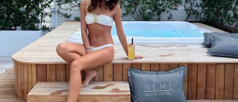 Una mujer sentada en una terraza de madera junto a una piscina, con una bebida, rodeada de follaje y una almohada del hotel WE.ME SUITE.