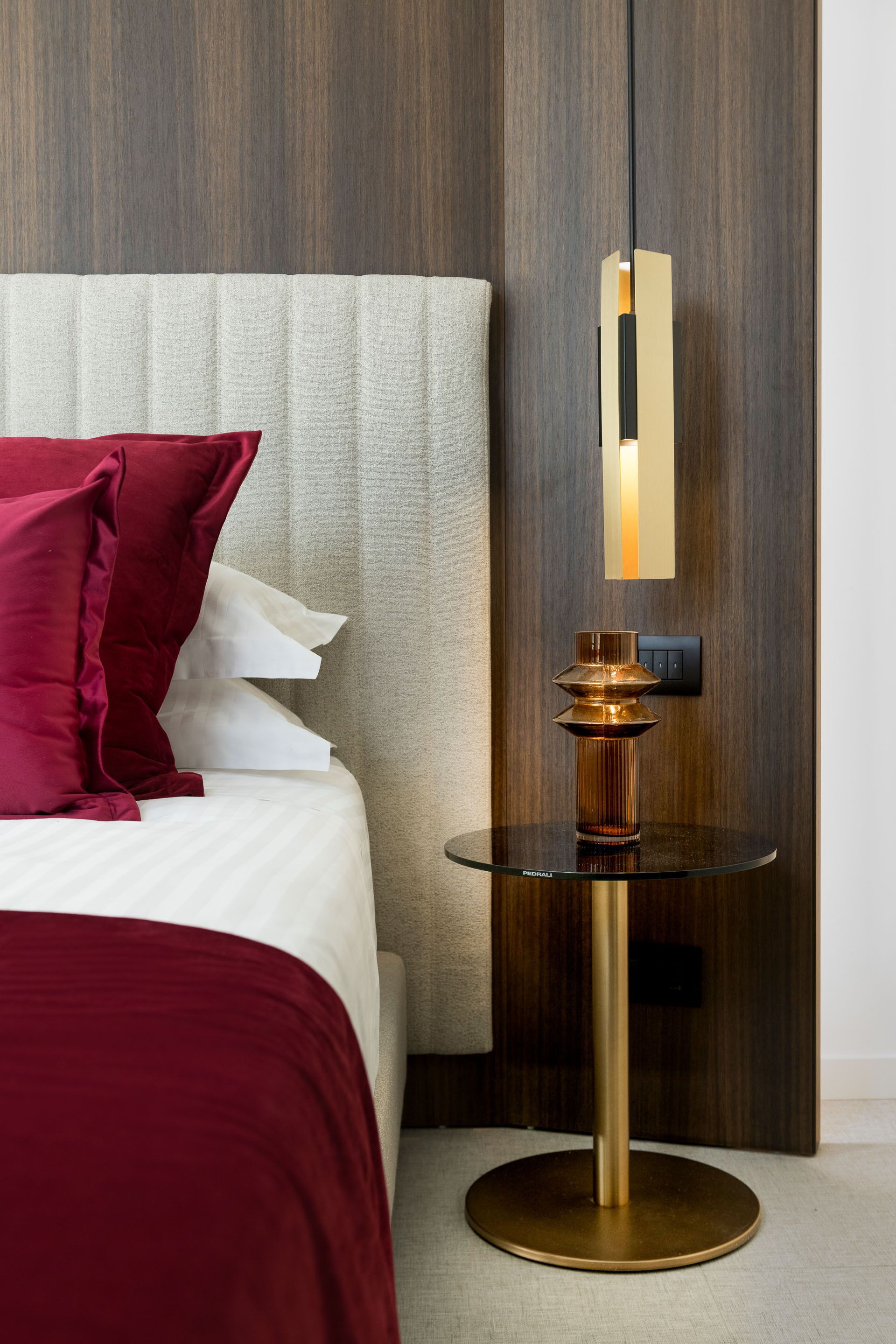 Una cama bien hecha con una colcha roja y blanca, una mesita de noche con marco dorado y un panel de pared moderno.