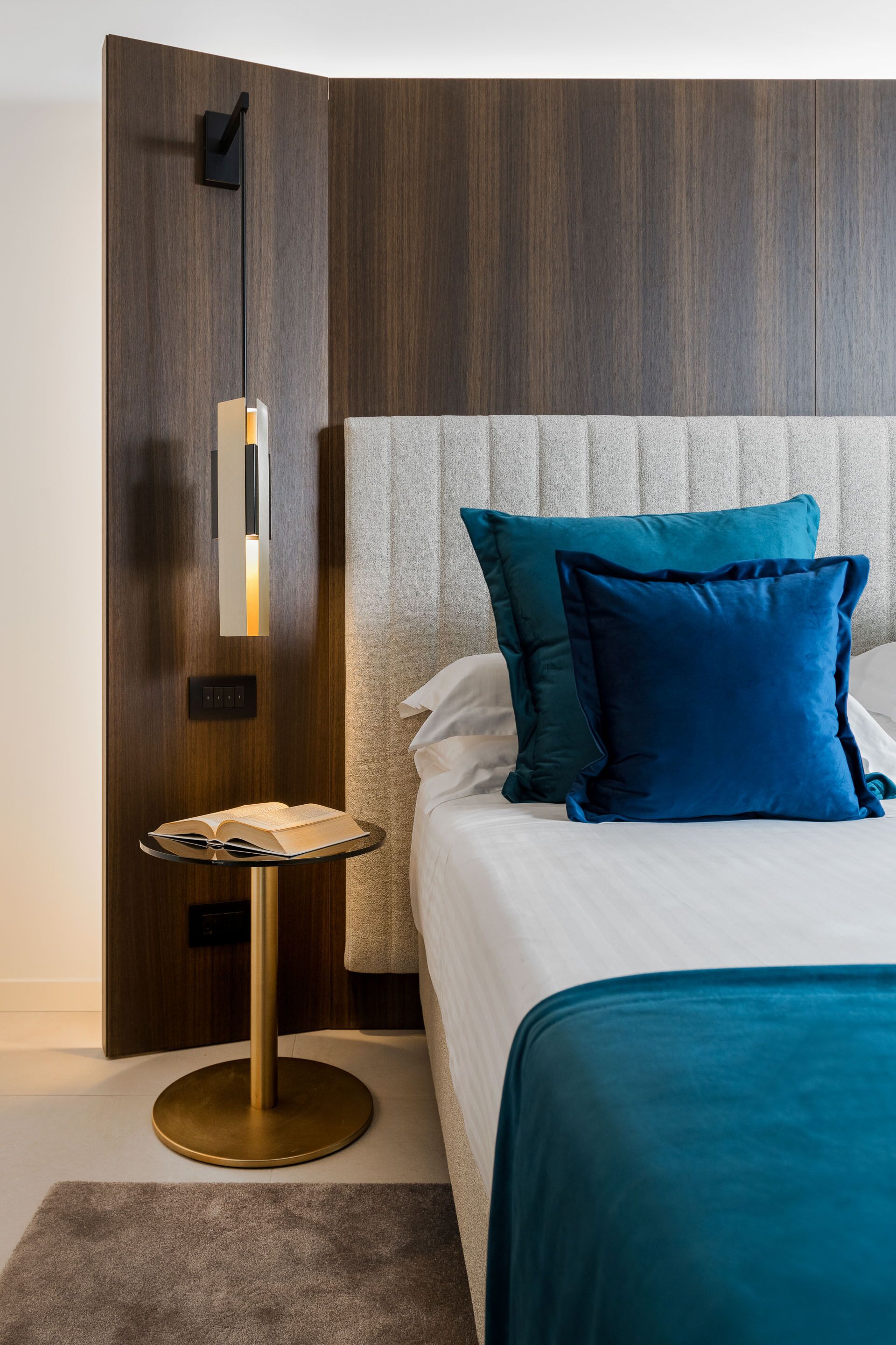 A neatly arranged hotel room with a bed, a bedside table with a book, and a modern wall panel.