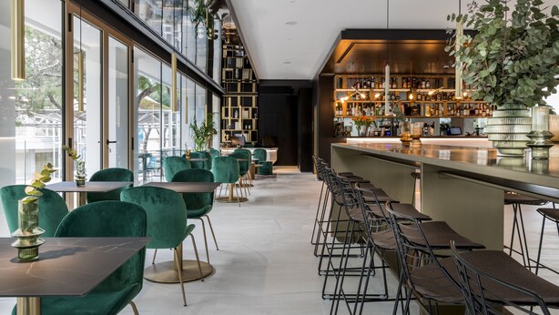 A modern bar area with green upholstered chairs, a long counter, and a variety of bottles and glasses. - We Me Suite Hotel (Riccione)