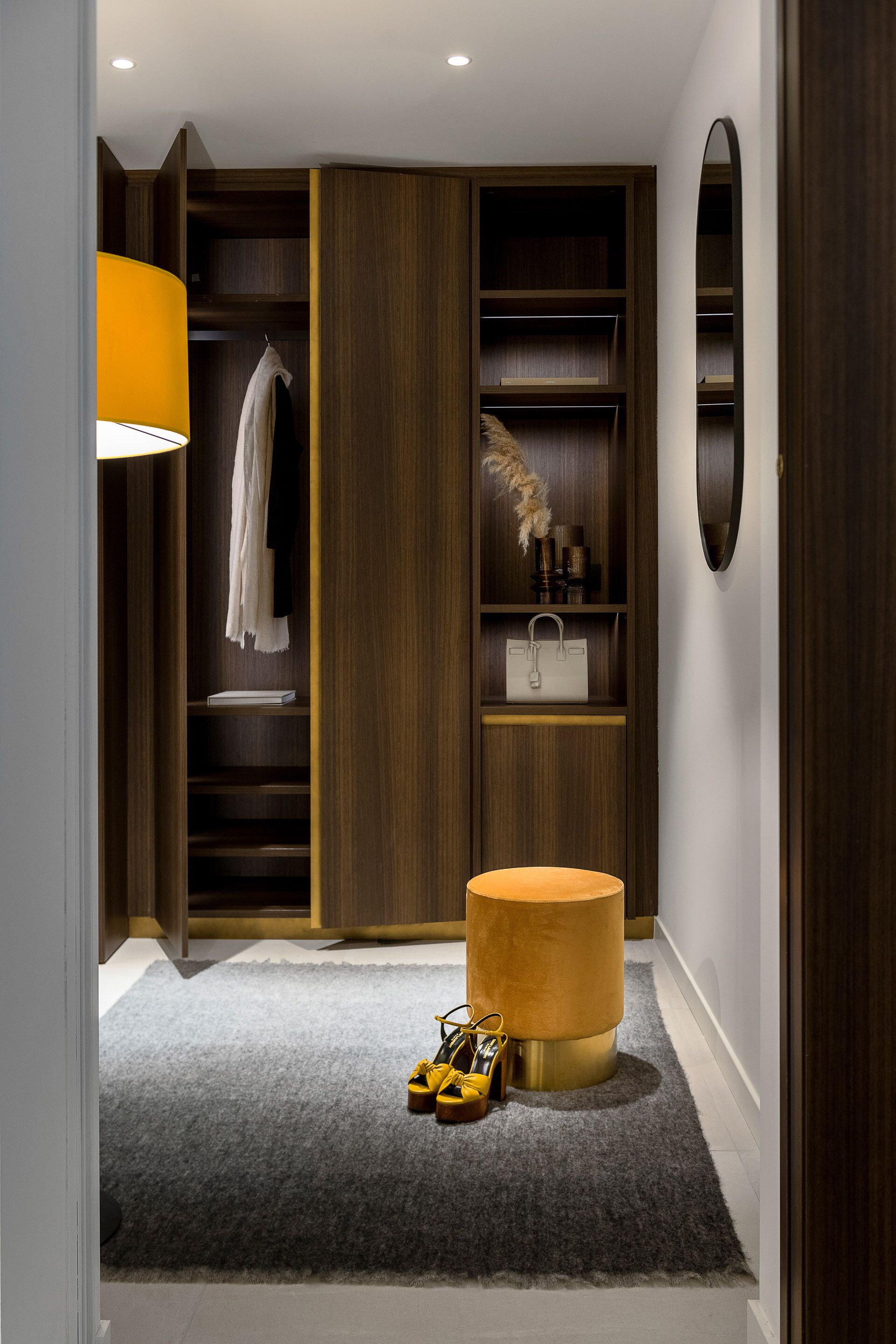 A modern walk-in closet with wooden shelving, a round ottoman, and a mirror.