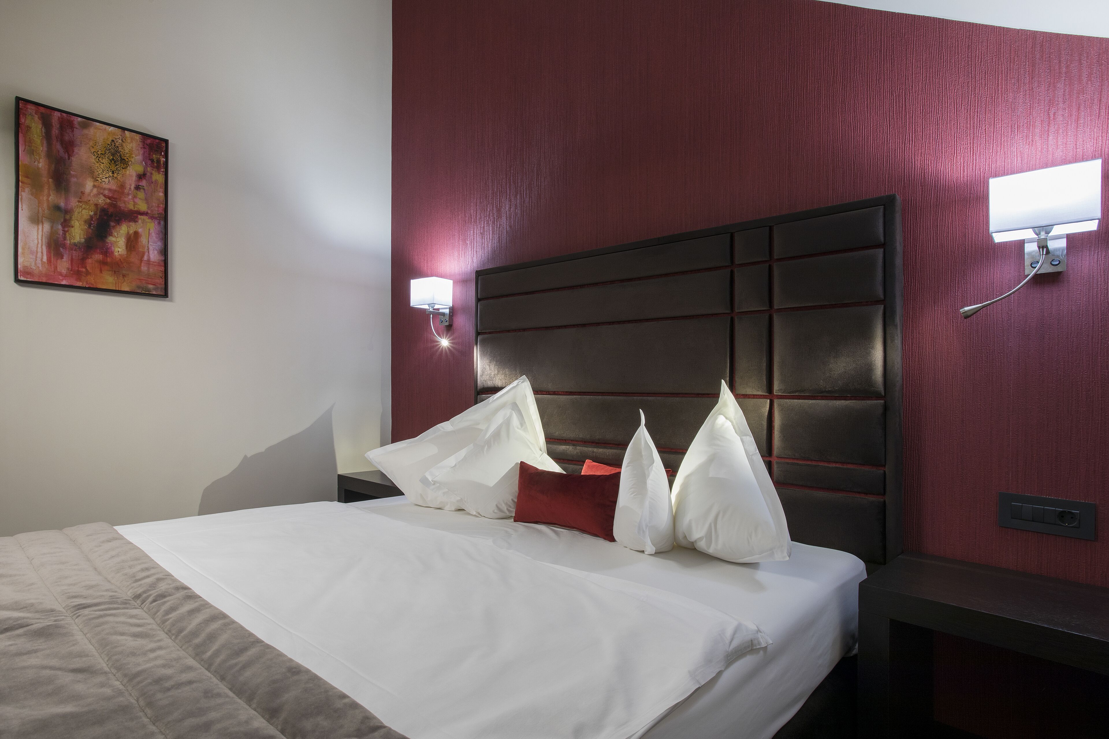 A hotel room with a large bed, a headboard with a panel design, a nightstand with a lamp, and a painting on the wall.