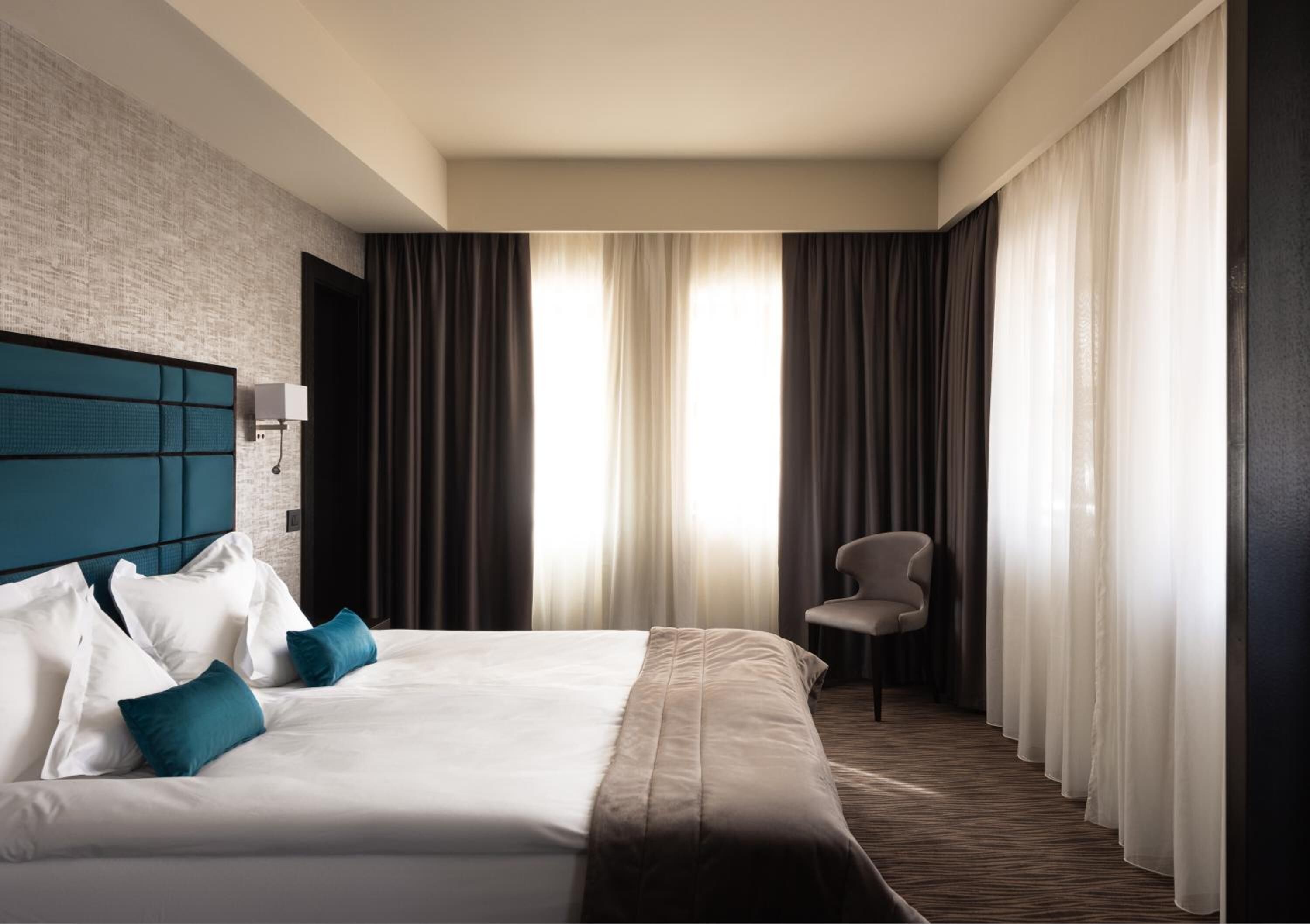 Une chambre d’hôtel avec un grand lit, une tête de lit, une chaise et une fenêtre avec des rideaux.