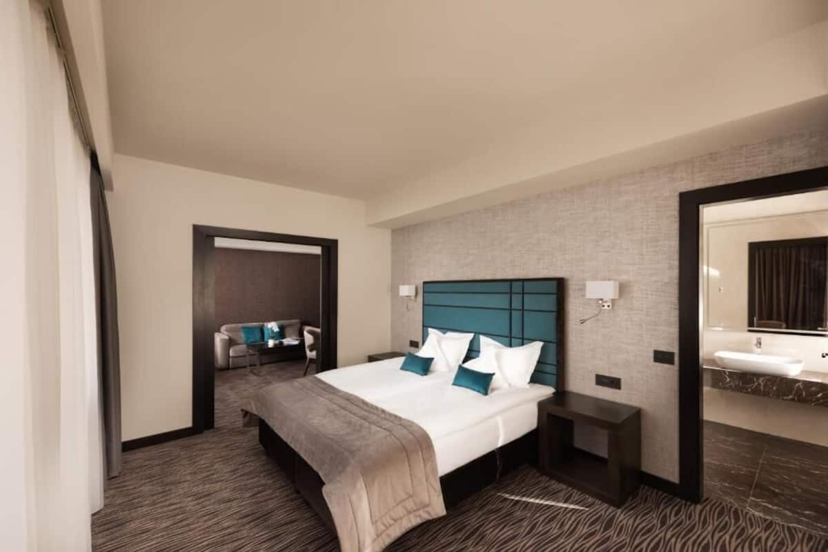 A hotel room with a large bed, a sitting area with a sofa and armchair, a bathroom with a mirror and sink, and a carpeted floor.