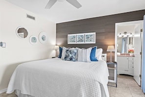 Une chambre à coucher comprenant un lit, des tables de chevet, une commode, un miroir, des œuvres d’art murales et un ventilateur de plafond.
