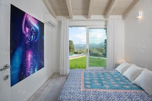 A bedroom with a large window, a bed with white and blue bedding, and a colorful abstract painting on the wall.