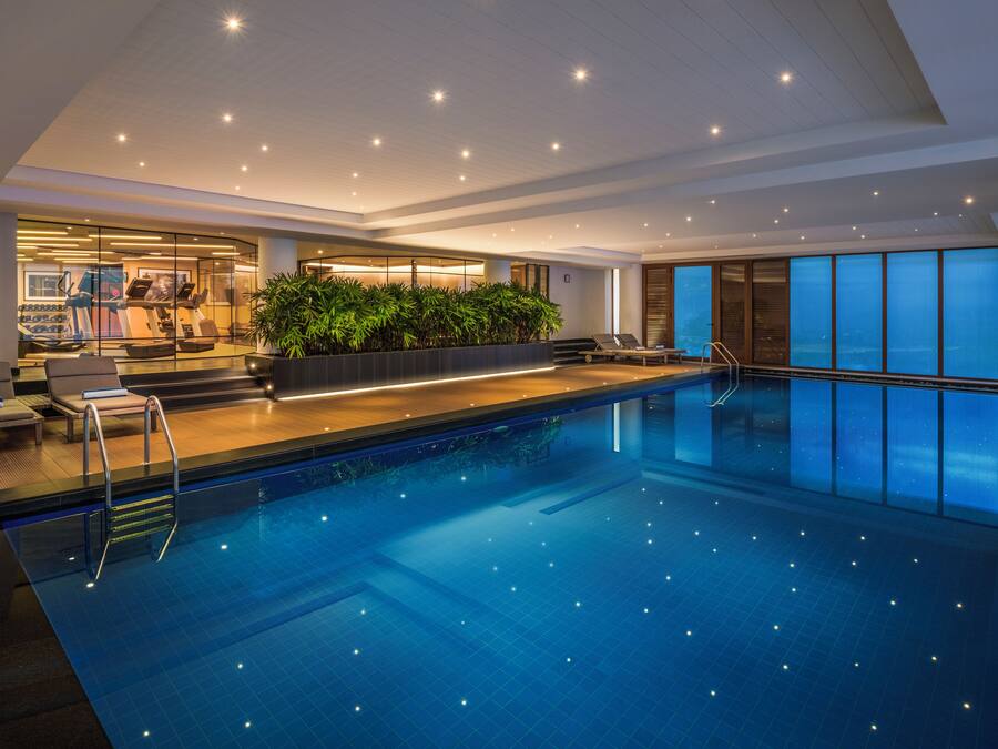 A modern indoor swimming pool with a glass wall, exercise area, and lounge chairs.