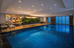 Una piscina interior moderna con pared de vidrio, zona de ejercicios y sillones de descanso.