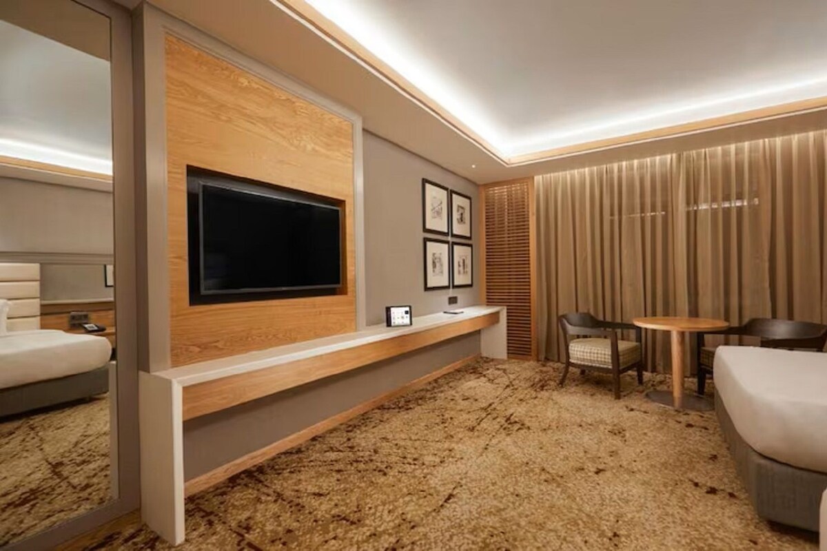 A hotel room with a TV mounted on a wooden panel, a small desk with a chair, a round table with two chairs, and a bed.