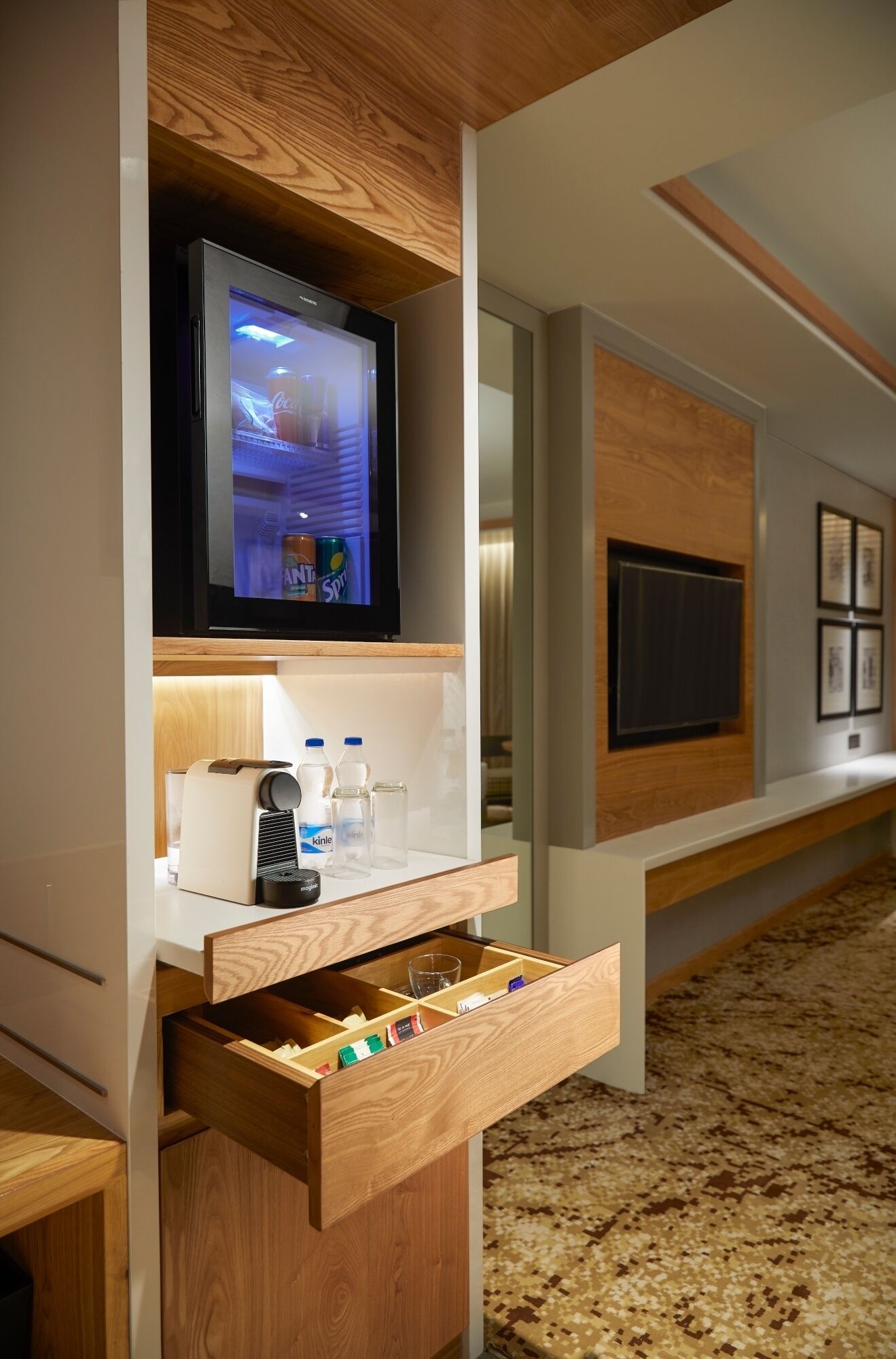 A modern hotel room with a built-in TV, minibar, and a wooden drawer unit.