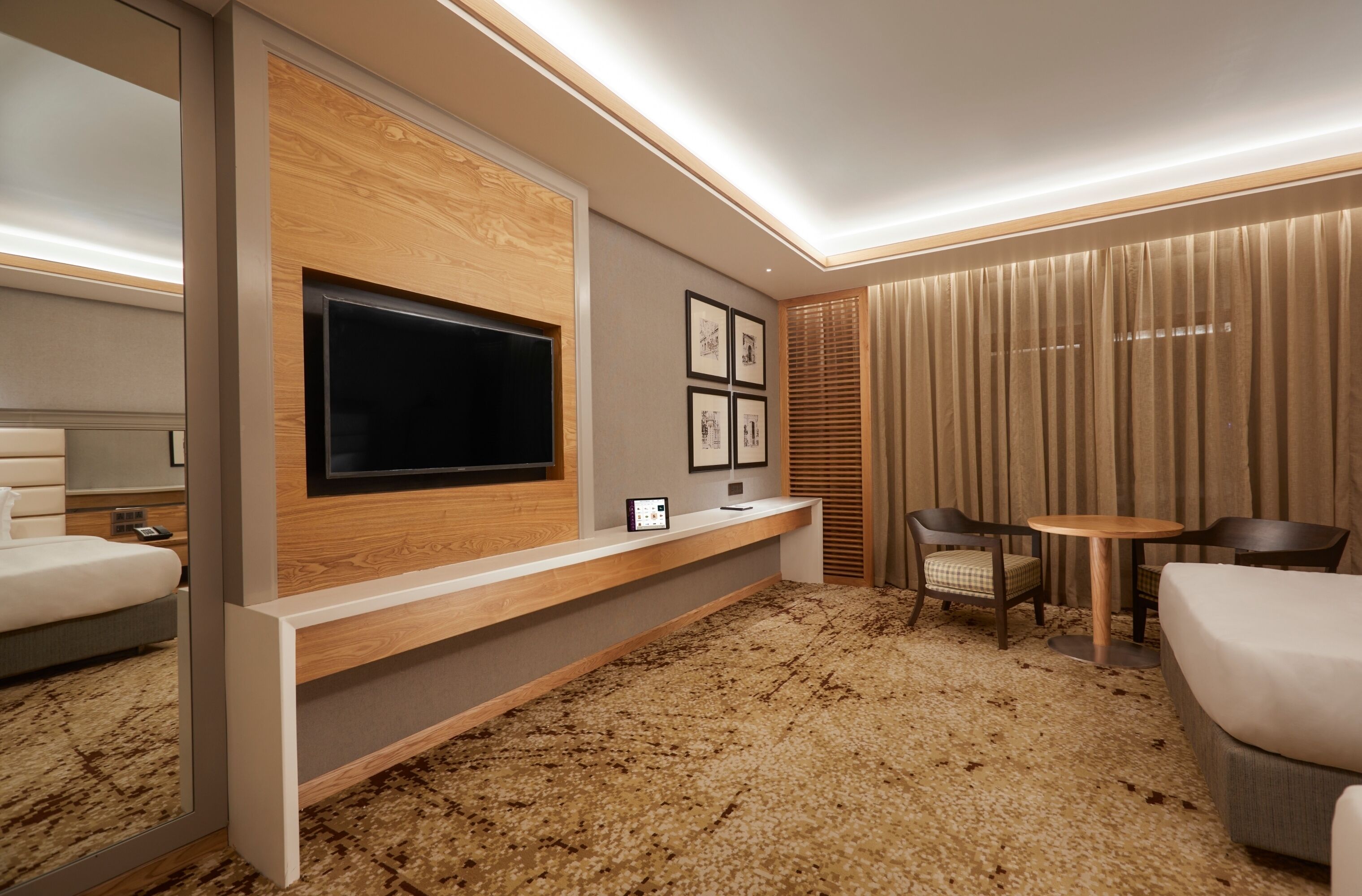 Ein modernes Hotelzimmer mit großem Flachbildfernseher an einer Holzvertäfelung, einem kleinen Tisch mit zwei Stühlen, einem Bett und einem Teppichboden.