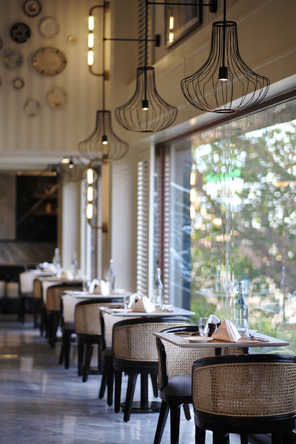 A restaurant interior with multiple tables and chairs, pendant lighting, and a view of greenery outside. - The Residency Towers Puducherry (Puducherry)