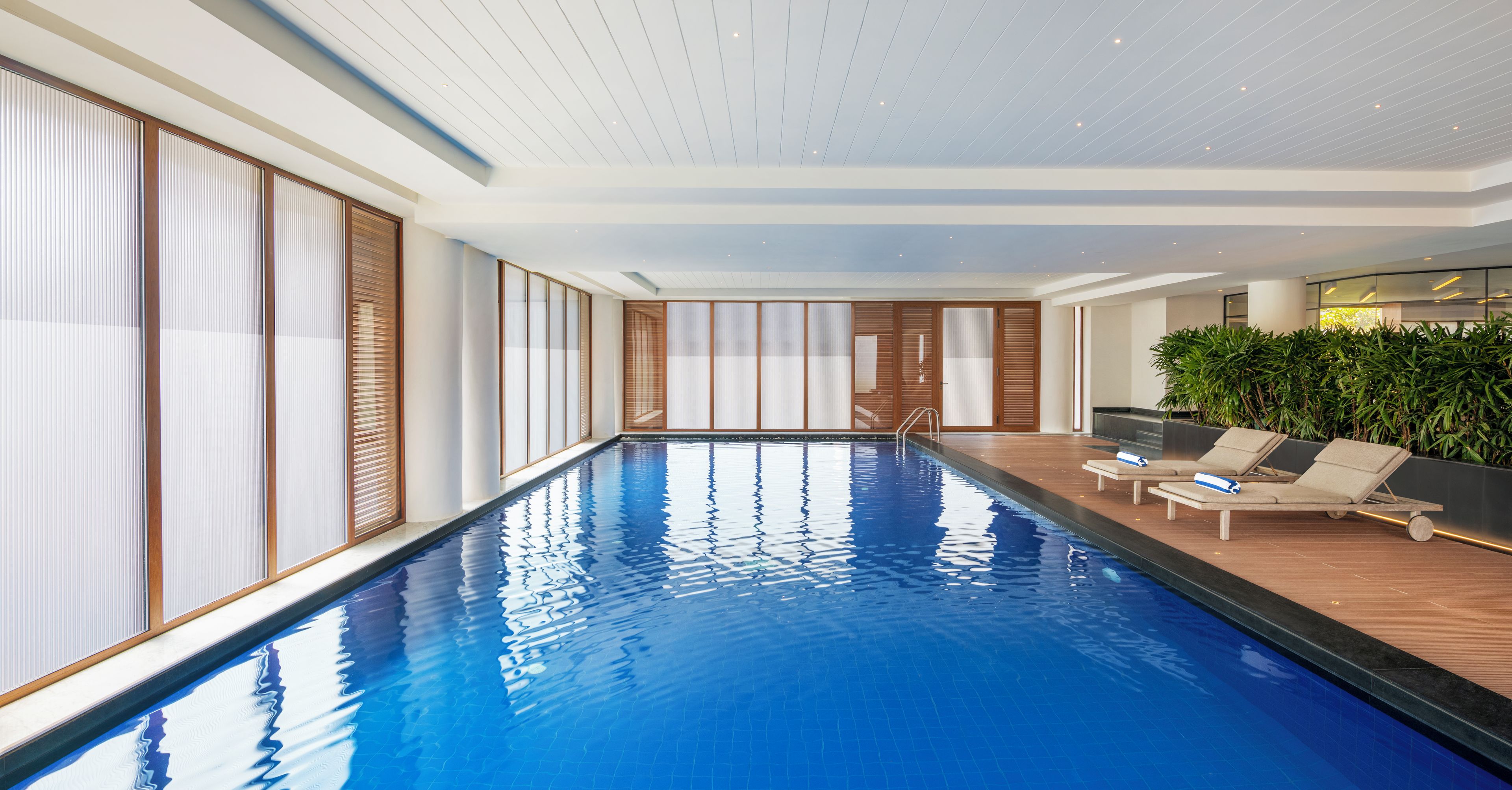 An indoor swimming pool with a wooden deck, lounge chairs, and a large window with blinds.