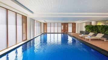 An indoor swimming pool with a wooden deck, lounge chairs, and a large window with blinds.
