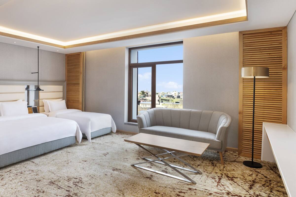 Une chambre d’hôtel moderne avec deux lits, un canapé, une petite table et une grande fenêtre donnant sur la ville.