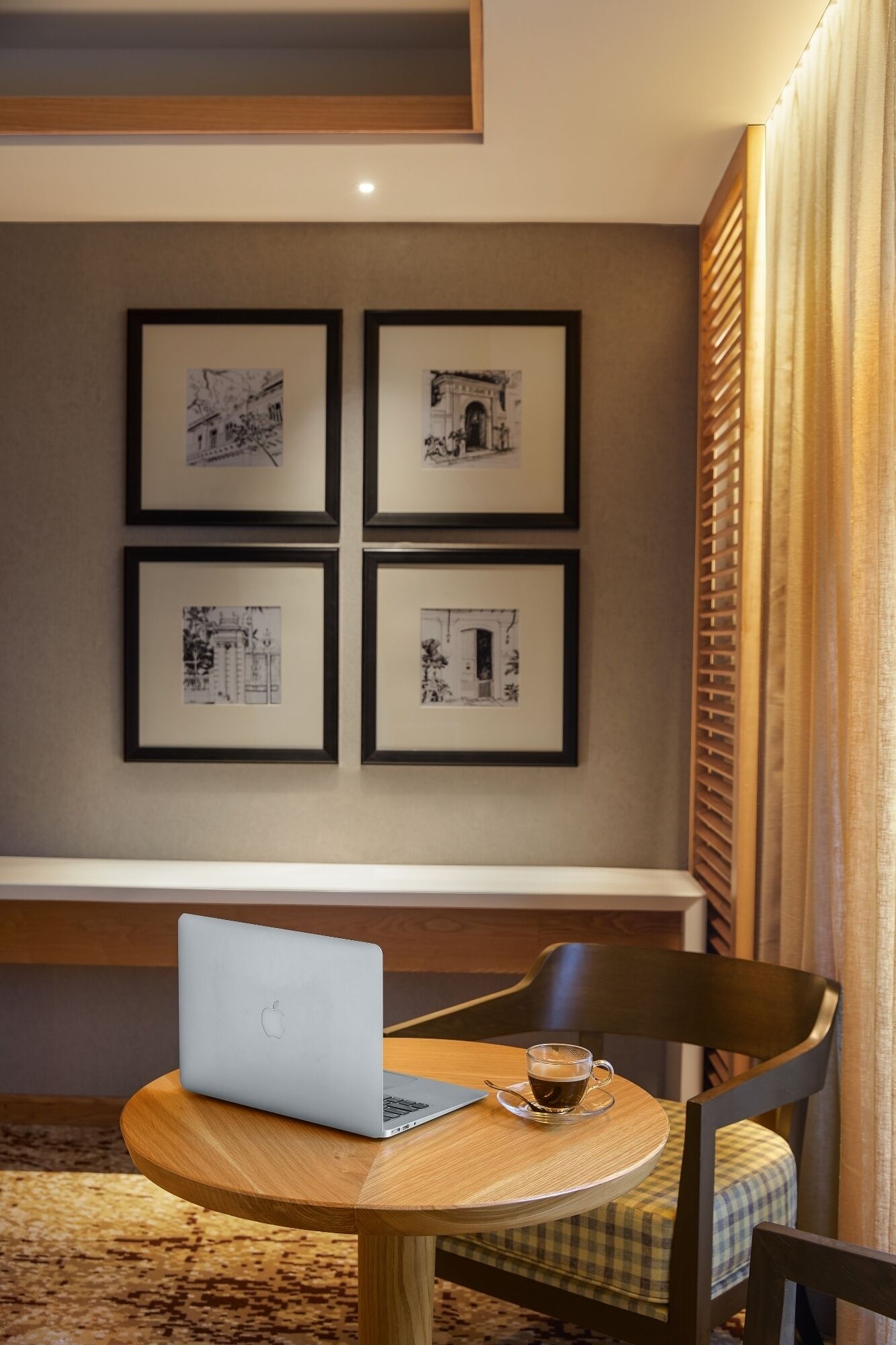 A hotel room with a wooden desk, a laptop, and a cup of coffee. There are four framed black and white photographs on the wall.