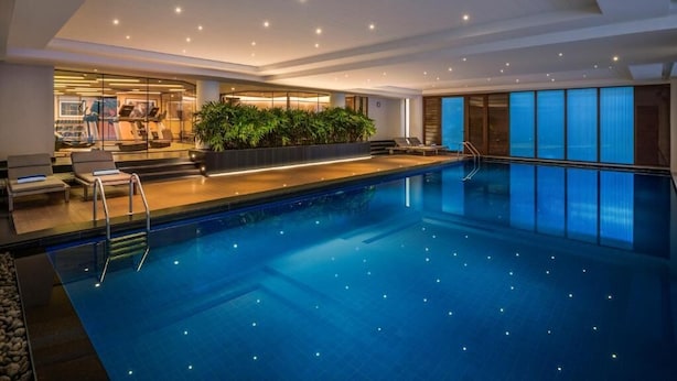 Piscina interior moderna com parede de vidro, cadeiras de descanso e área de ginástica.