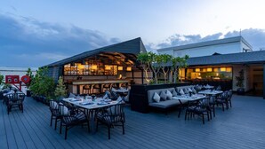 Uma área de refeições ao ar livre com mesas e cadeiras, um espaço de bar com assentos e um edifício moderno com telhado inclinado.