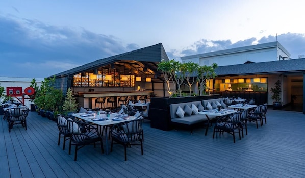 Uma área de refeições ao ar livre com mesas e cadeiras, um espaço de bar com assentos e um edifício moderno com telhado inclinado.