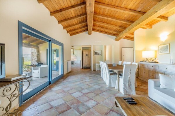 A spacious living area with a high wooden ceiling, a dining table with chairs, a sofa, and a TV on a stand.