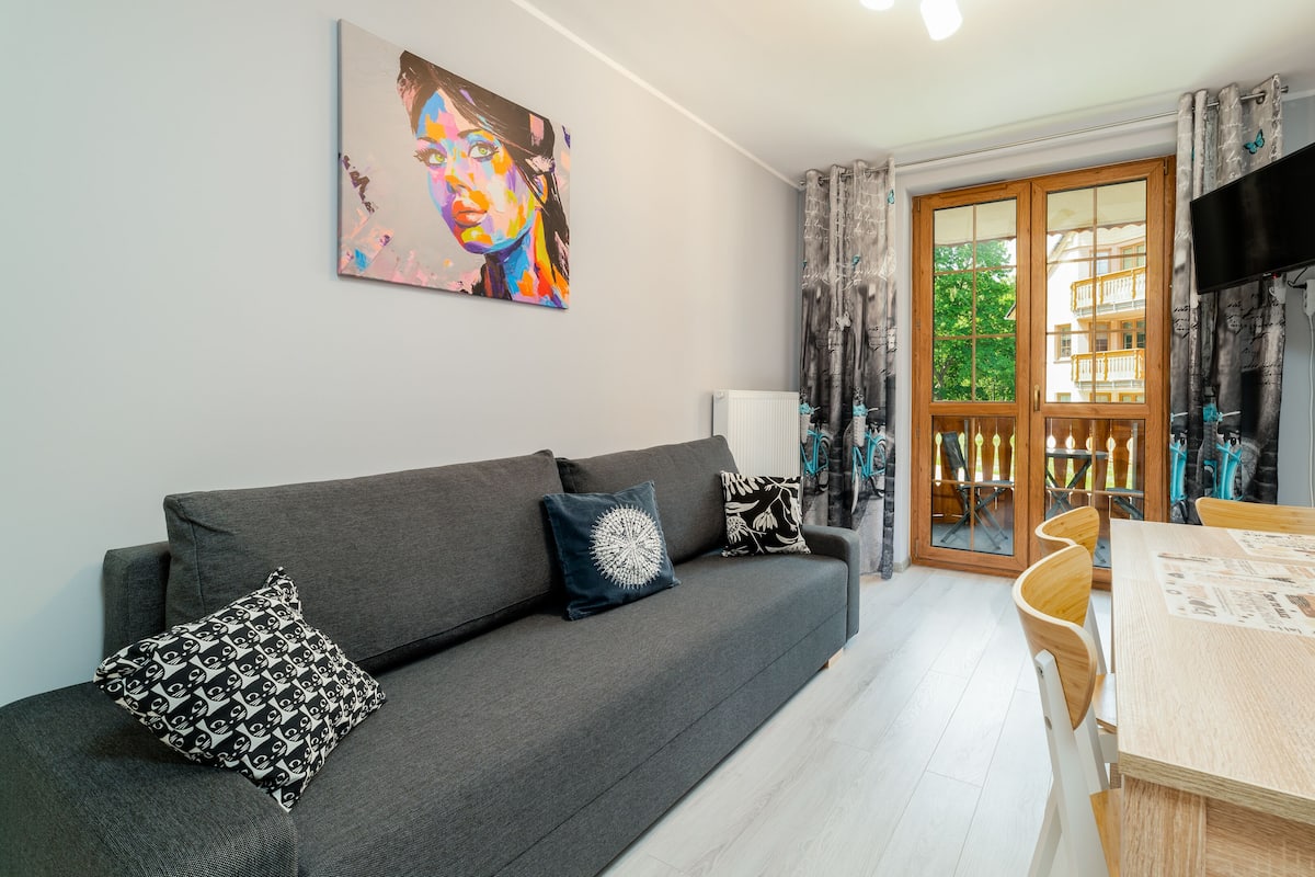 Ein modernes Wohnzimmer mit grauem Sofa, zwei gemusterten Kissen, einem Esstisch mit Stühlen, einem an der Wand montierten Flachbildfernseher und Blick auf einen Balkon mit Gartenmöbeln.