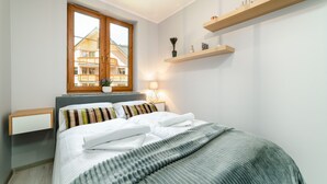 Een slaapkamer met een bed, een nachtkastje, een raam met uitzicht op gebouwen, een lamp en houten schappen.