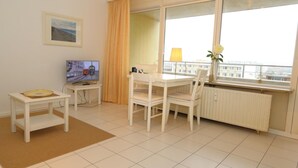 A hotel room with a dining table, chairs, a TV, a radiator, and a window with a view of buildings. - Metropol House, apt. 109 (Sylt)