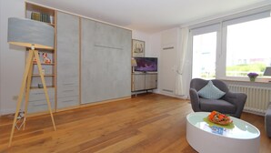 A modern living room with a wooden floor, a grey sofa, a coffee table with flowers, a TV, and a wardrobe with a mirror. - App. Kohfahl 65, Germany (Sylt)
