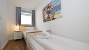A single bed with white linens, a bedside table with a lamp, a window with curtains, and a painting of a beach scene on the wall. - App. Kohfahl 65, Germany (Sylt)