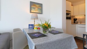 A dining area with a table set for two, a painting on the wall, and a kitchen in the background. - App. Kohfahl 65, Germany (Sylt)